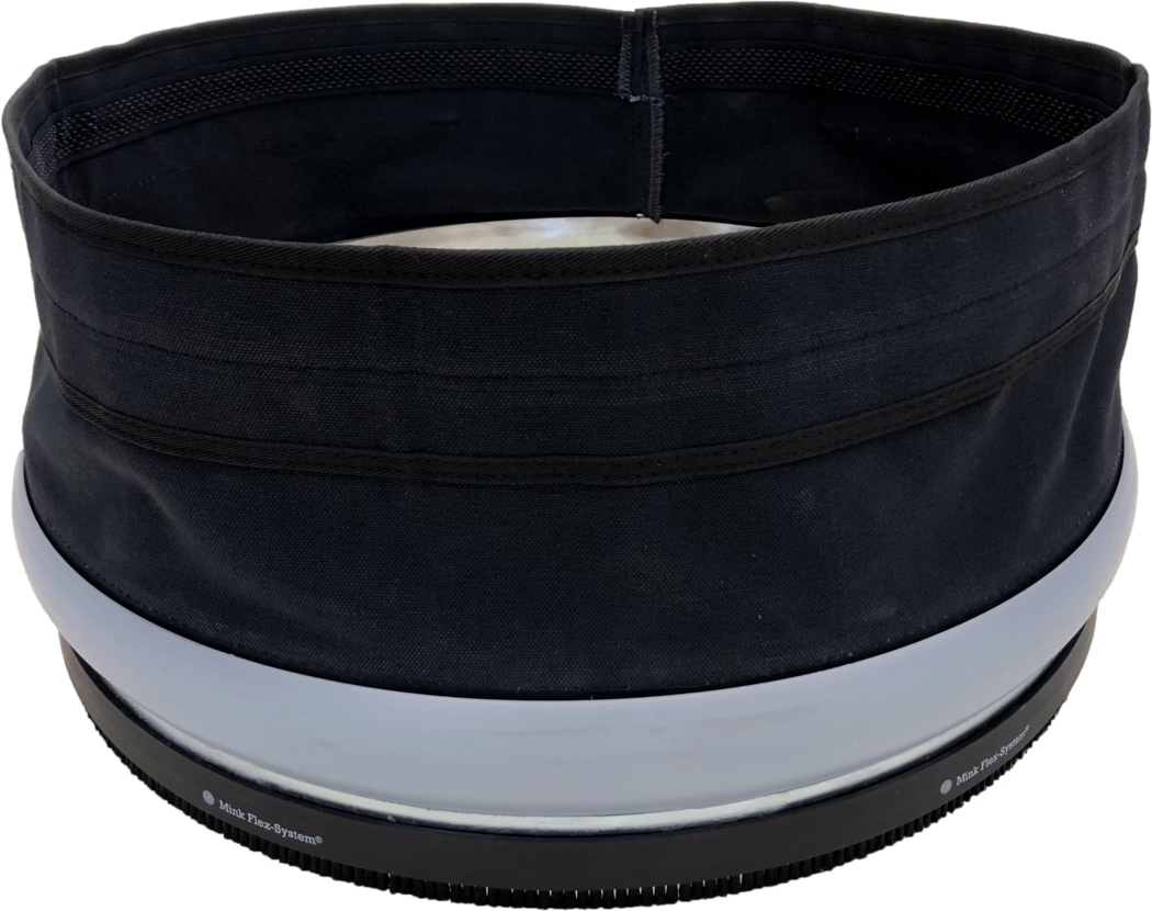 Gros plan du bord supérieur et de l'ouverture d'un contenant cylindrique en tissu noir ou d'un chapeau. La photo montre une ouverture circulaire intérieure avec une doublure noire, des coutures renforcées visibles et plusieurs bandes horizontales en tissu à l'extérieur. Près du bord inférieur se trouvent deux larges anneaux en plastique gris clair et une base en caoutchouc noir striée avec de petites dents et un texte imprimé partiellement visible 'Mink Flex-System®'. Le tissu présente un tissage grossier et une finition mate; l'objet paraît structuré mais souple. L'éclairage est uniforme et l'arrière-plan est blanc.