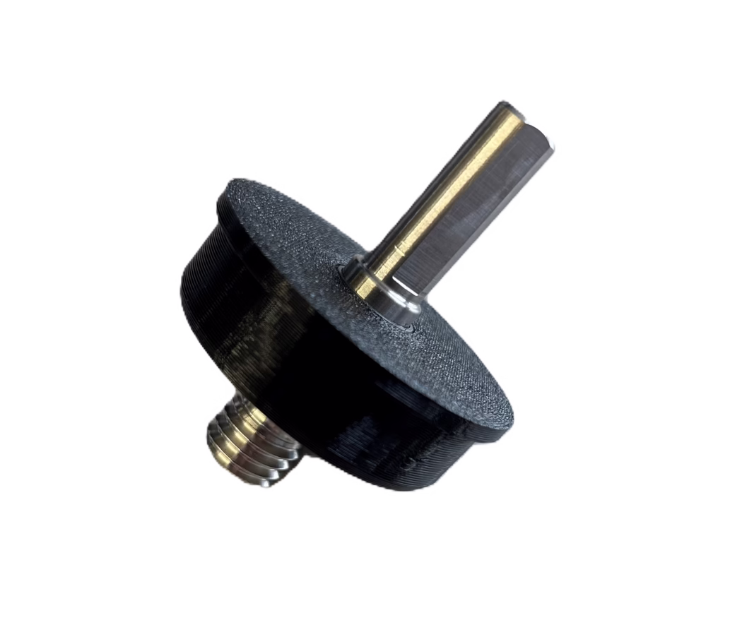 PALLMANN_product image_adapter cordless screwdriver Gros plan d'un petit ensemble mécanique : un arbre métallique hexagonal avec un revêtement couleur laiton inséré dans un bouton ou un entretoise en plastique noir ou imprimé en 3D. L'arbre métallique présente une face plate polie et des traces d'usinage visibles ; une tige filetée métallique avec des filets concentriques est visible à la base. La pièce noire montre une surface texturée et striée typique de l'impression 3D FDM. L'objet est photographié en diagonale sur un fond blanc propre.